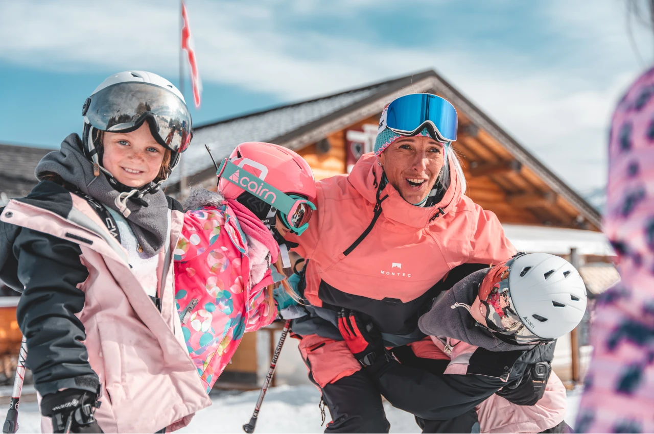 Un moniteur de ski avec 3 enfants devant une cabane en bois dans la neige